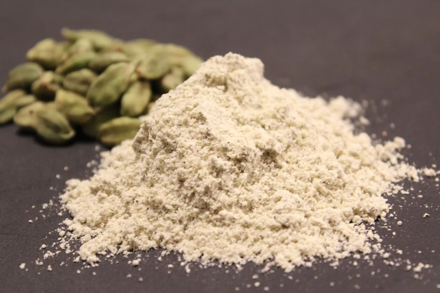 Un mucchietto di cardamomo in polvere su un piano scuro, con capsule verdi intere sullo sfondo