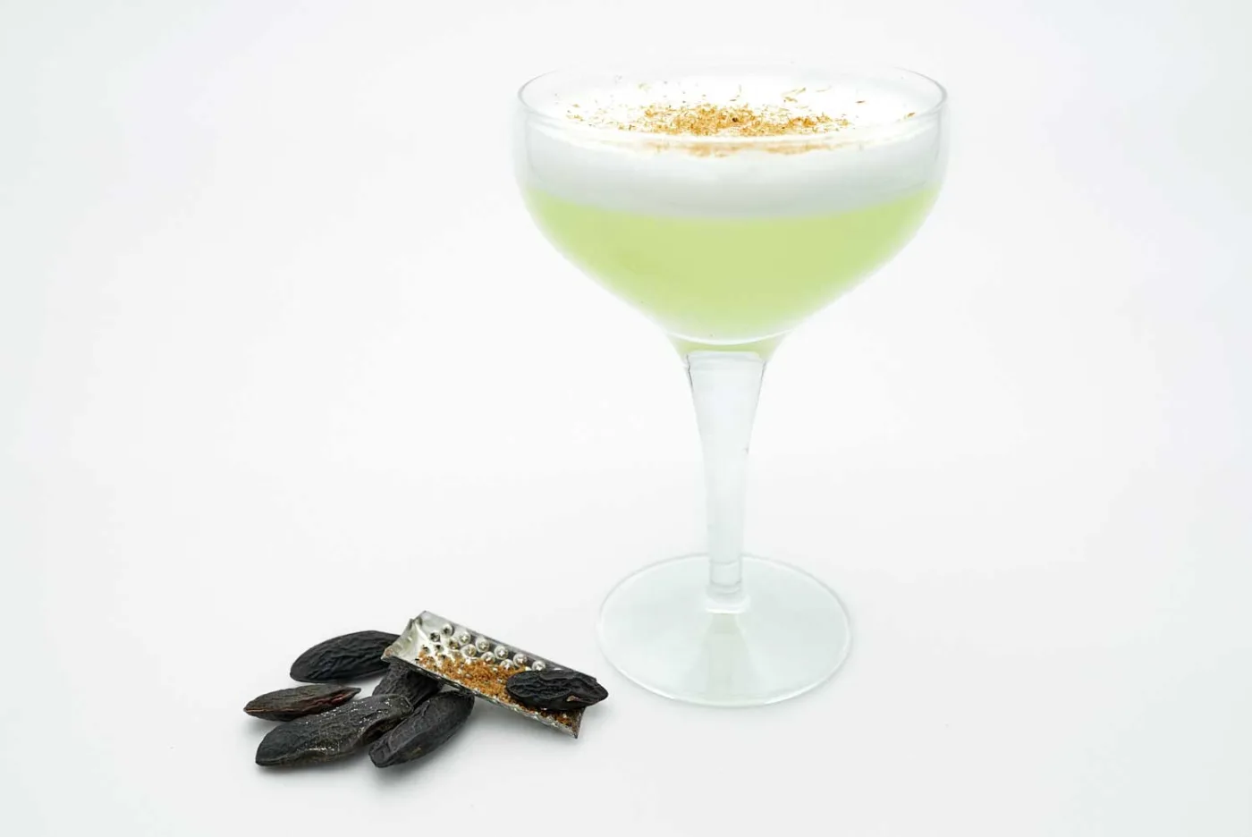 Un Inca Sour in coppetta, verde chiaro con schiuma bianca e fava di tonka grattugiata sopra, accanto a una piccola grattugia e alcune fave