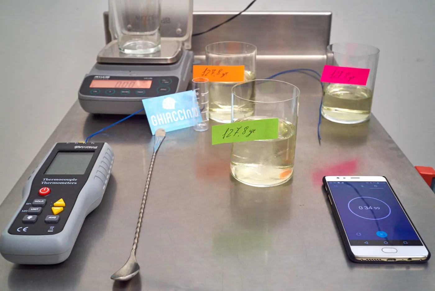 Postazione dell’esperimento con termometro a termocoppia, bar spoon, cronometro sullo smartphone e i bicchieri etichettati