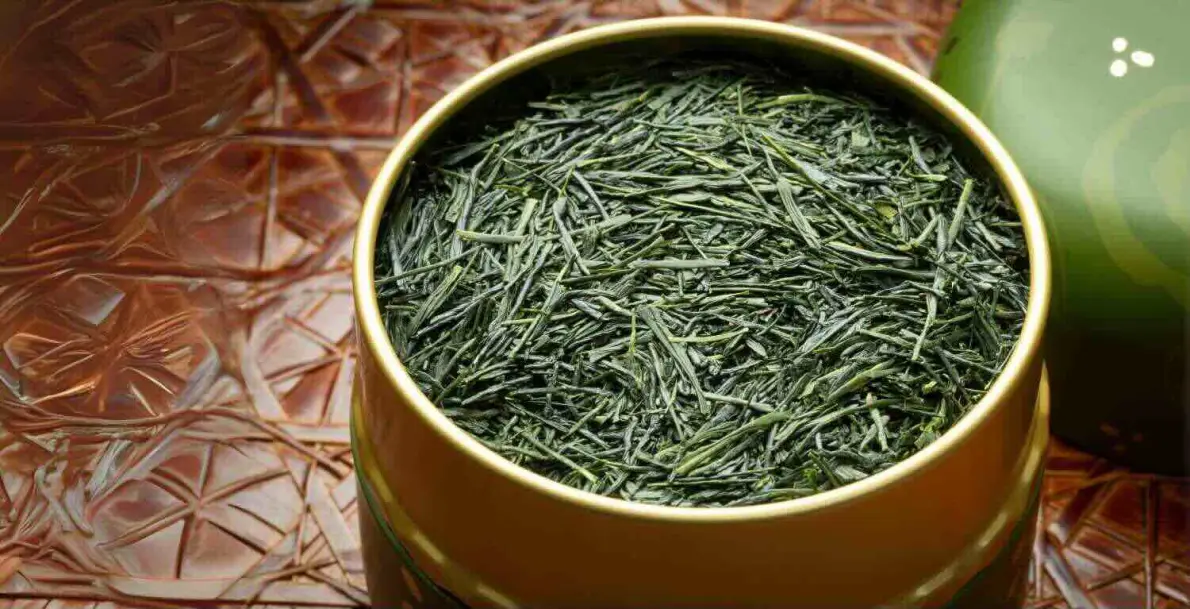 Una scatola rotonda dorata piena di foglie di tè Gyokuro verde scuro, su un panno rosso