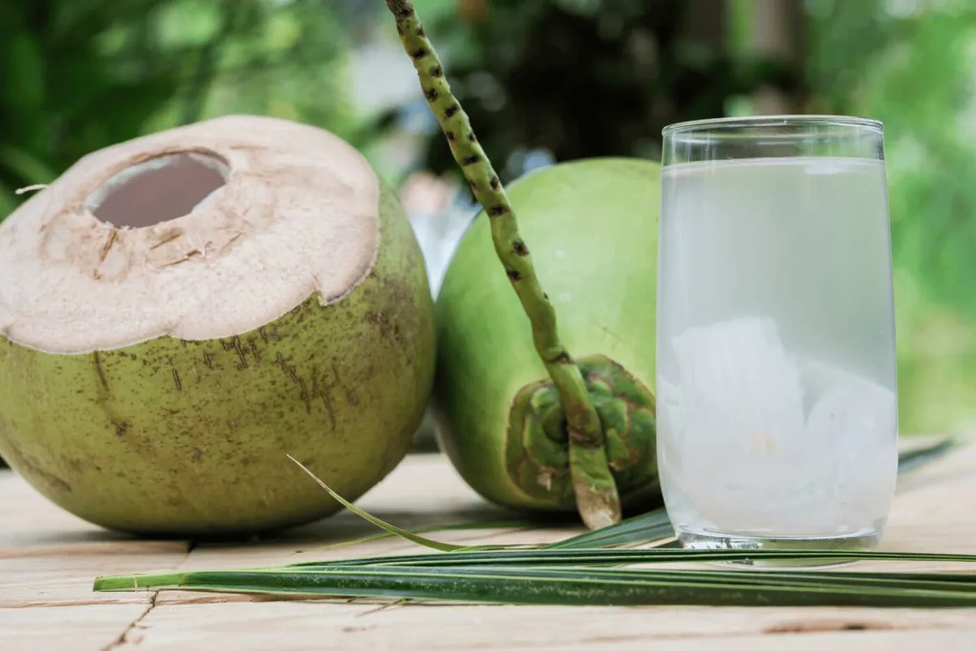 Due cocchi verdi, uno aperto in cima, accanto a un bicchiere di acqua di cocco su un tavolo di legno