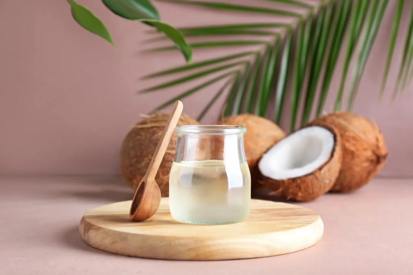 Un vasetto di vetro con olio di cocco e un cucchiaio di legno su un tagliere rotondo, con noci di cocco e una foglia di palma sullo sfondo