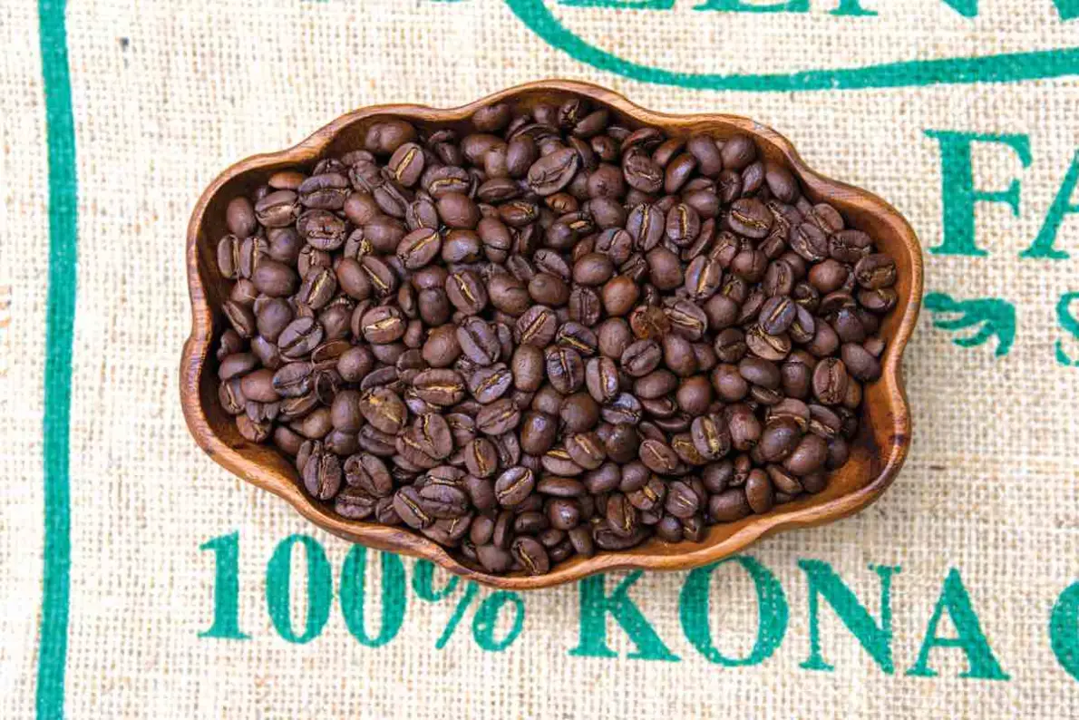 Chicchi di caffè Kona tostati in una ciotola di legno su un sacco di juta con la scritta 100% Kona