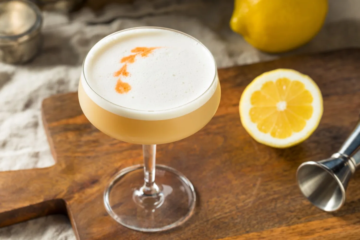 Cocktail sour in coppa con schiuma bianca e gocce di bitters in superficie, accanto a mezzo limone e un jigger