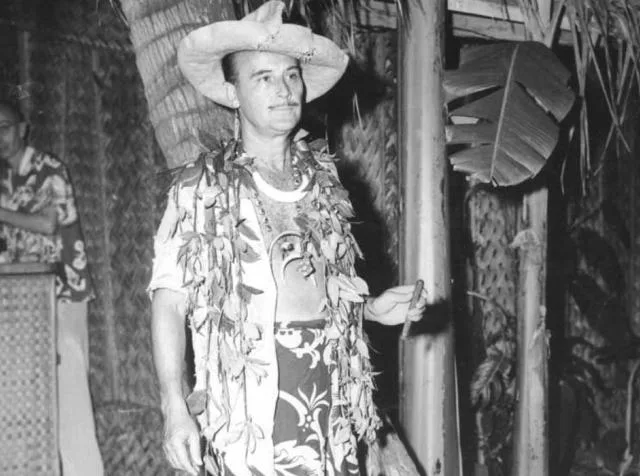 Fotografia in bianco e nero di Donn Beach con cappello di paglia, camicia hawaiana e collana di fiori davanti a una parete di bambù