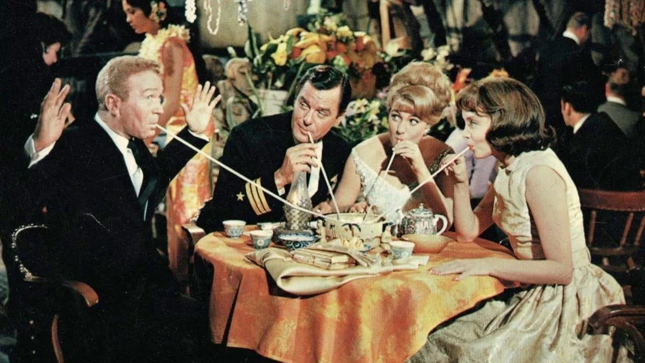 Quattro attori bevono con lunghe cannucce da un unico recipiente al centro del tavolo, in una scena del film Le astuzie della vedova (1963)