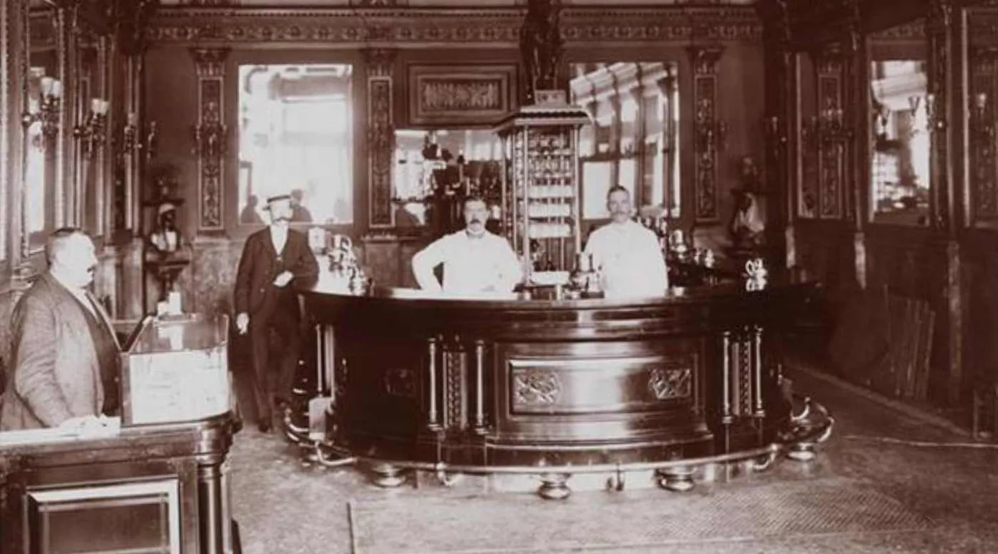 Fotografia seppia del bancone semicircolare dell'Hoffman House Bar con due barman in giacca bianca e un cliente