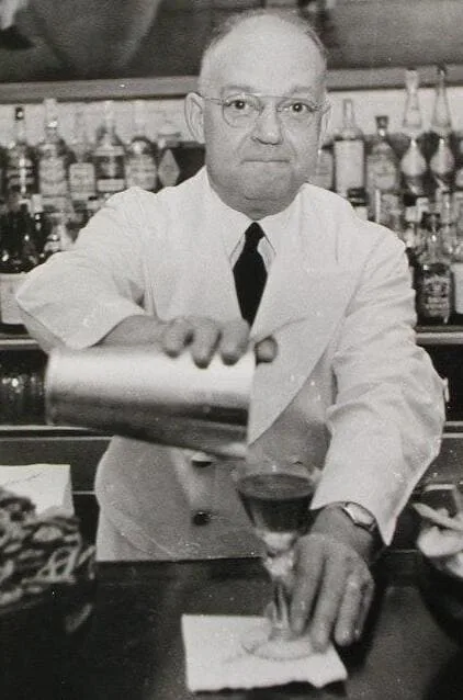 Fotografia in bianco e nero di Fernand Petiot, in giacca bianca e occhiali, mentre versa da uno shaker in una coppetta al bancone del bar