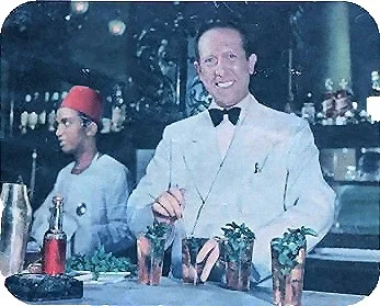 Fotografia a colori di Joe Scialom in giacca bianca e papillon dietro il bancone, mentre prepara drink decorati con la menta, con un cameriere con il fez alle spalle