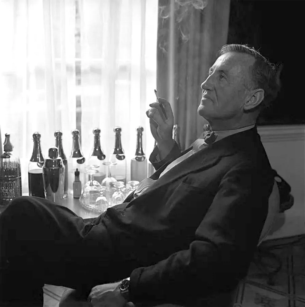 Ian Fleming seduto con una sigaretta in mano, in bianco e nero, con bottiglie e uno shaker sul tavolo dietro di lui